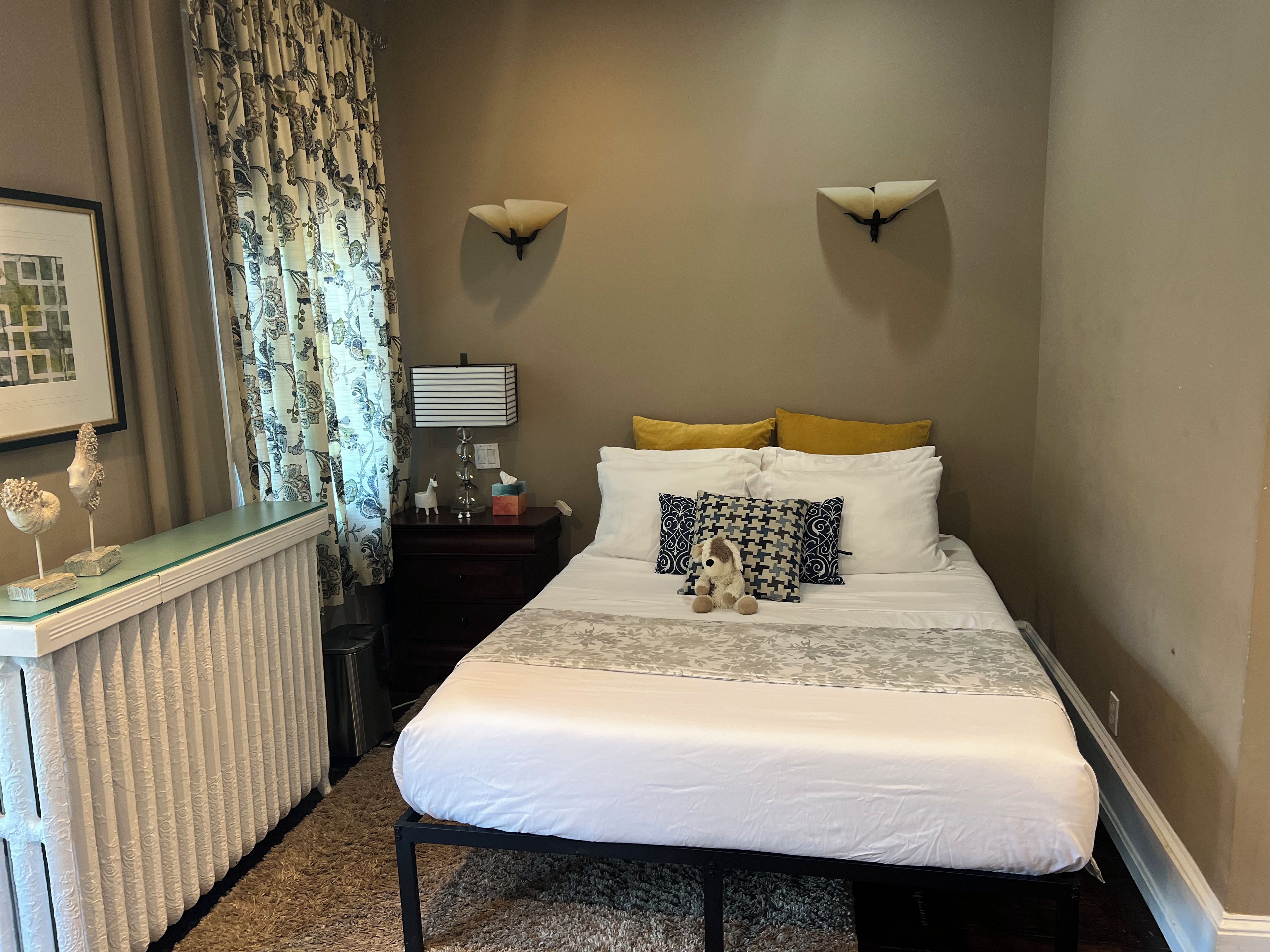 A neatly made bed with white bedding, a small bedside table with a lamp, and a wall-mounted light fixture.