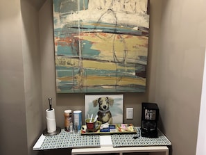 Un baño con un cuadro abstracto grande, un retrato enmarcado de un perro y una cafetera sobre una mesa con diseño.