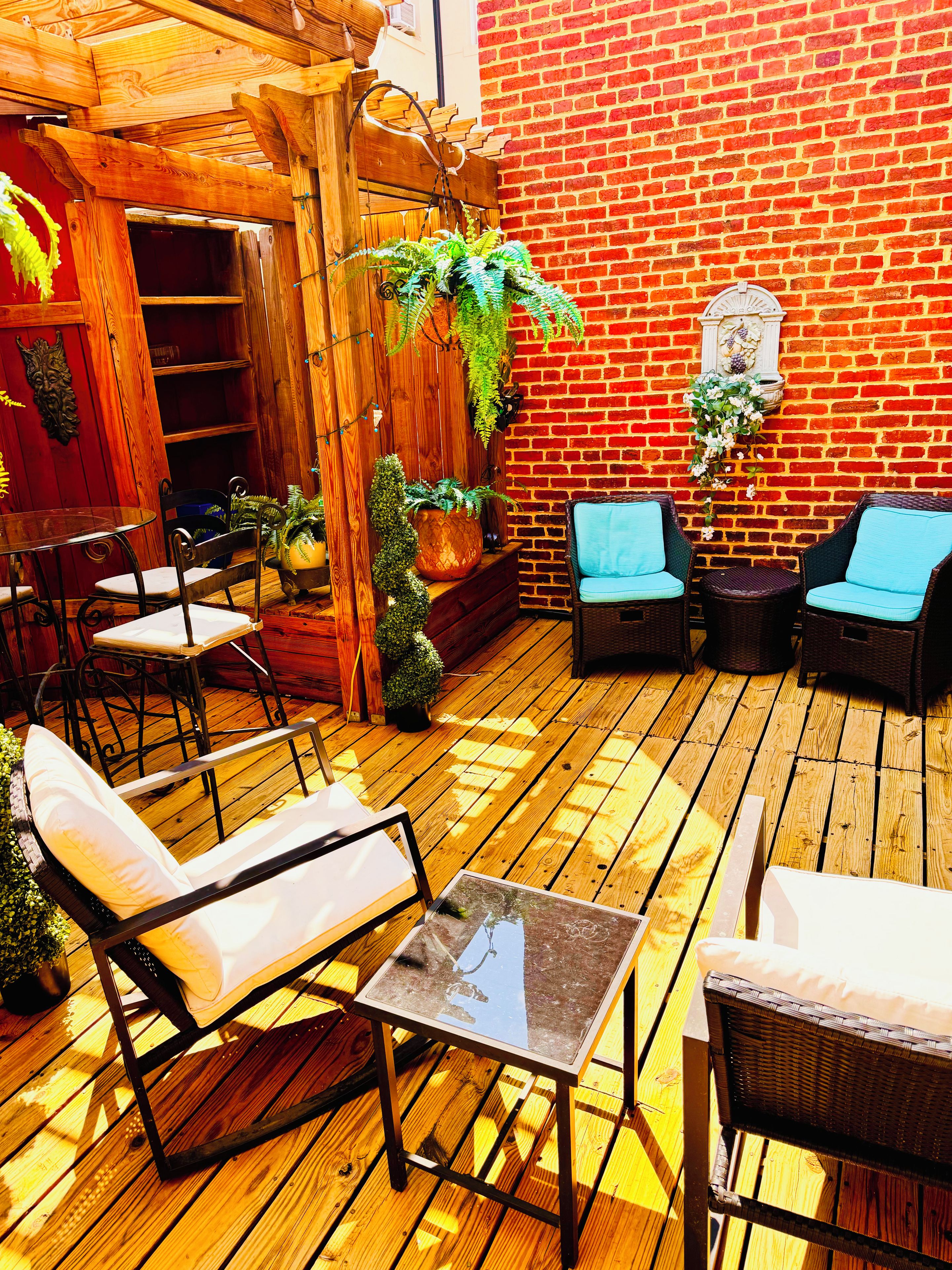 A wooden deck with outdoor furniture, including chairs and a table, surrounded by potted plants and a brick wall.