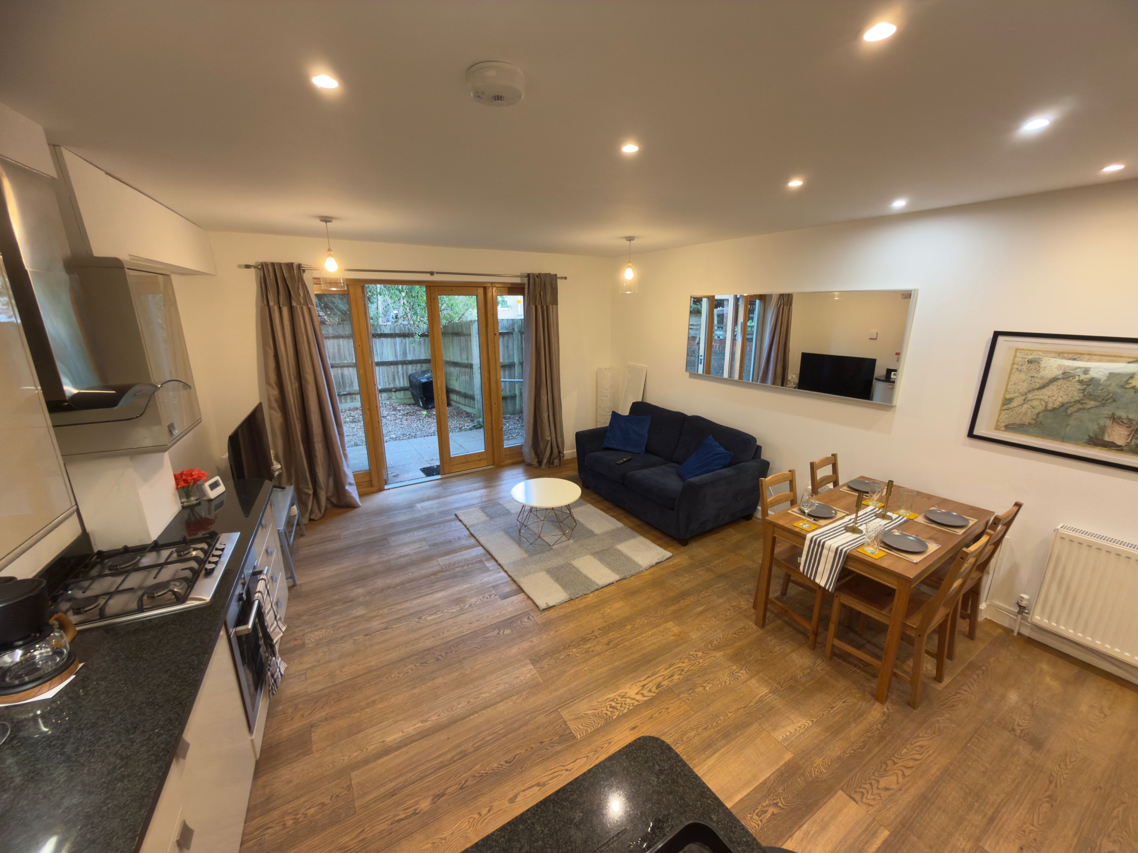 A modern kitchen with a gas stove, a dining area with a table set for two, a blue sofa, and a sliding glass door leading to a backyard.