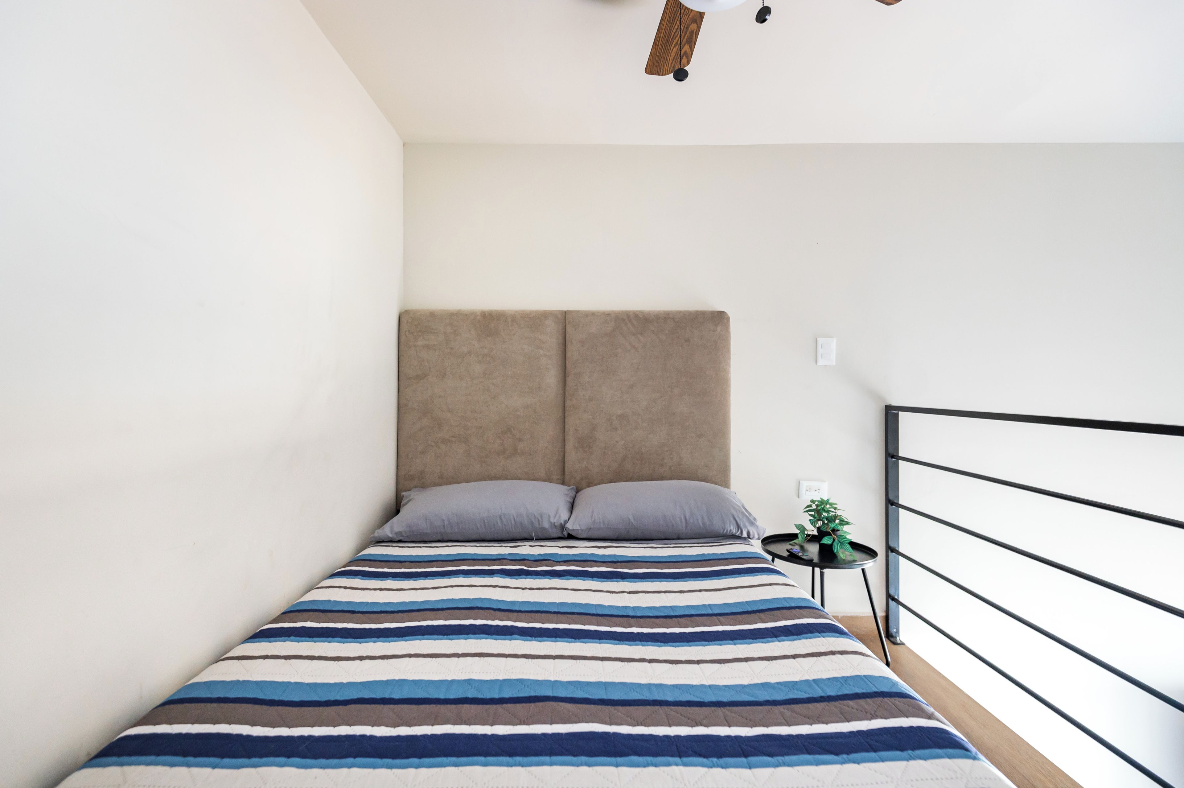 A bedroom with a bed, a headboard, a bedside table with a plant, and a railing.