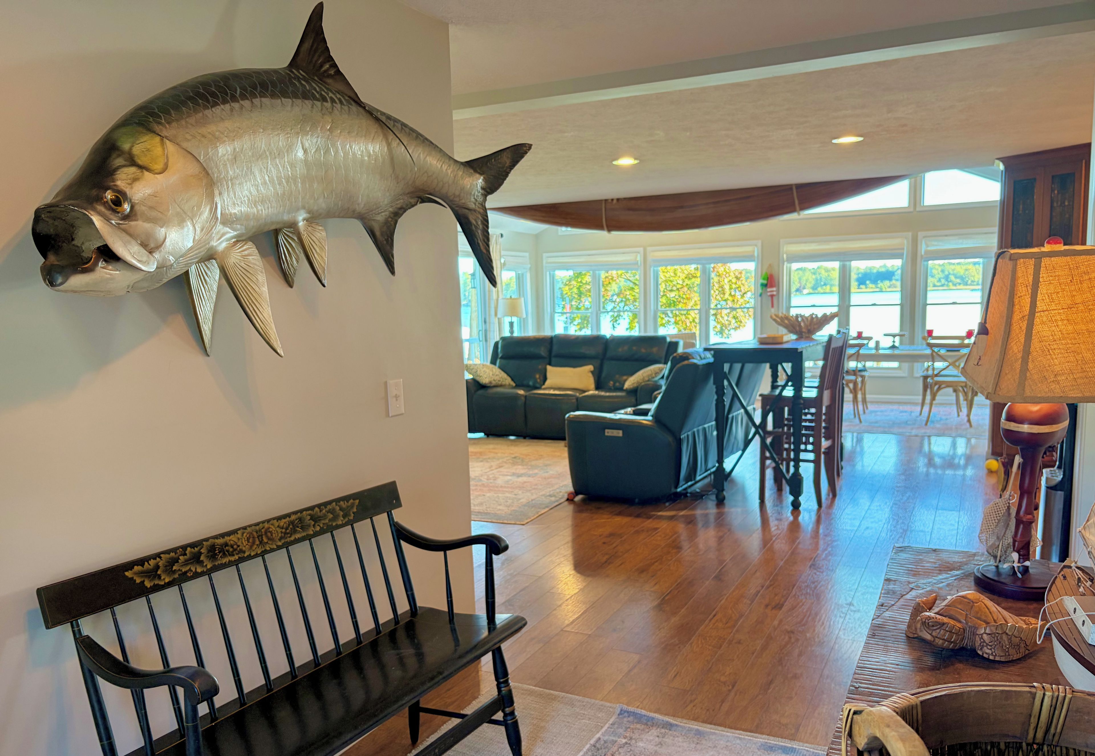 A room with a mounted fish on the wall, a bench, a sofa, a dining table with chairs, and a view through large windows.