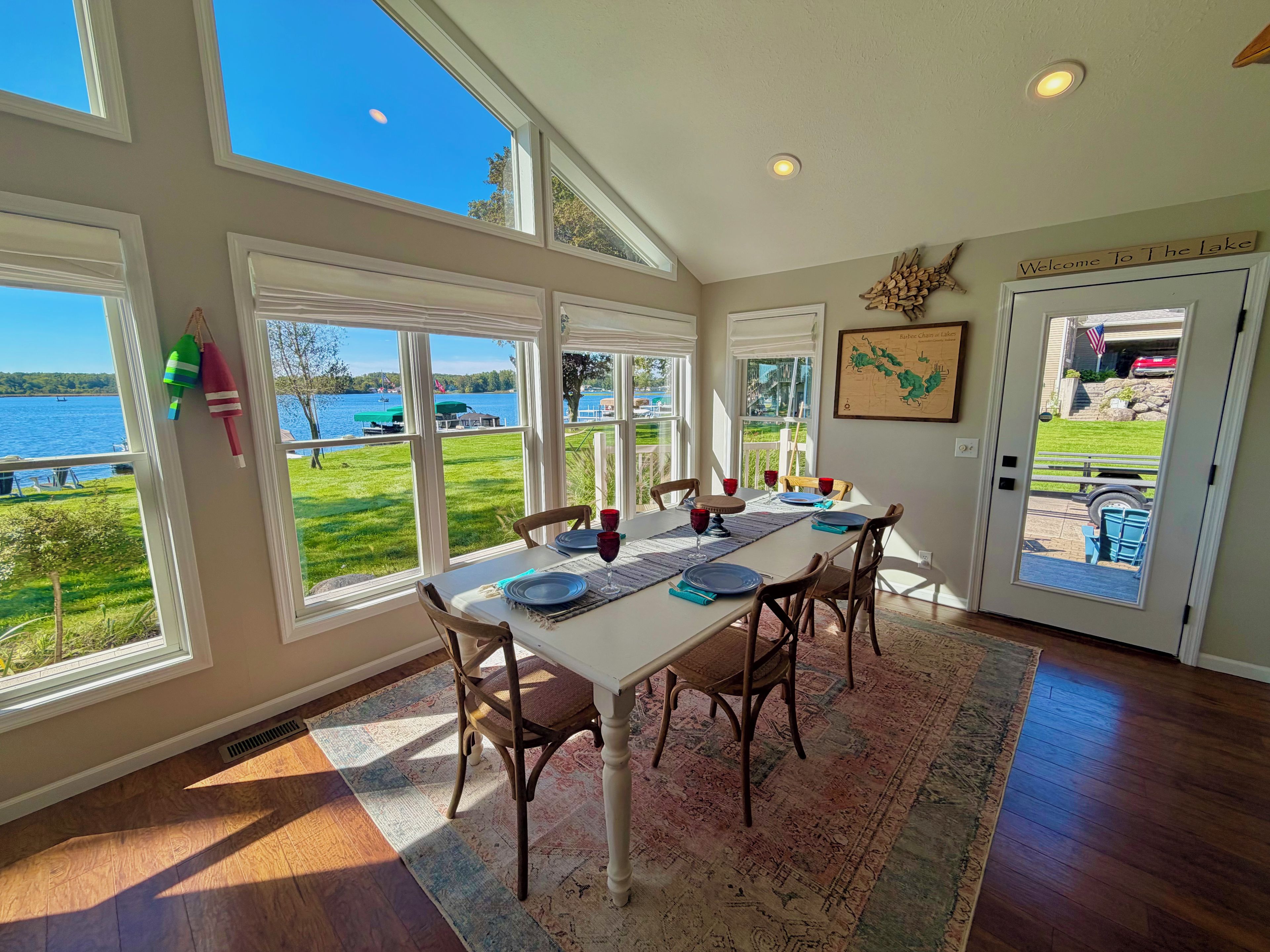 A dining area with a table set for four, large windows offering a view of a lake, and a door with a welcome sign.