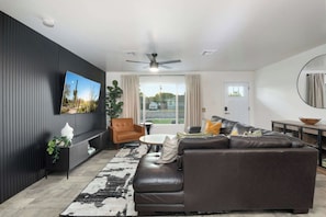 A modern living room with a sectional sofa, a television mounted on the wall, a ceiling fan, and a large window with curtains. - NEW| The Tipsy Cactus Old Town Oasis|Pool+Bar+Putt (Scottsdale)