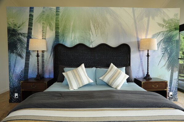 Una cama con cabecera oscura, dos mesitas de noche con lámparas y un mural temático tropical.