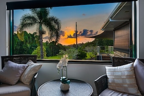 Una habitación con vista a un paisaje al atardecer, un sillón de mimbre, una mesita con un florero y una ventana con vista a una casa y vegetación.