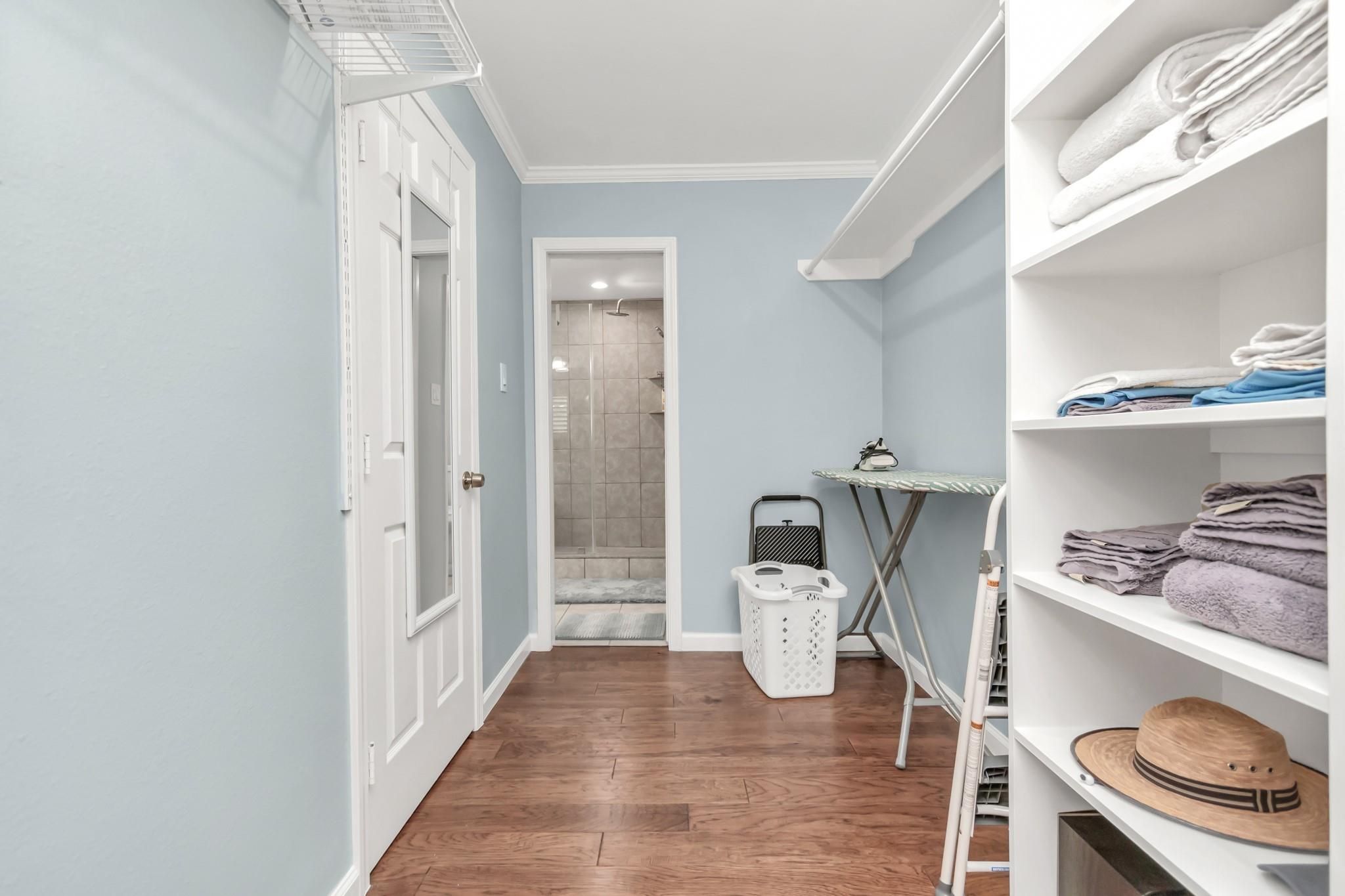 Un couloir avec un plancher en bois, un coin buanderie avec un tableau à repasser, un panier et des étagères remplies de vêtements pliés.