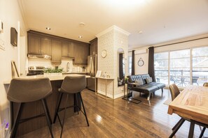 A modern kitchen with wooden cabinets, a stainless steel refrigerator, and a bar area with stools. - Loft Doré | Montmorency Falls (Boischatel)