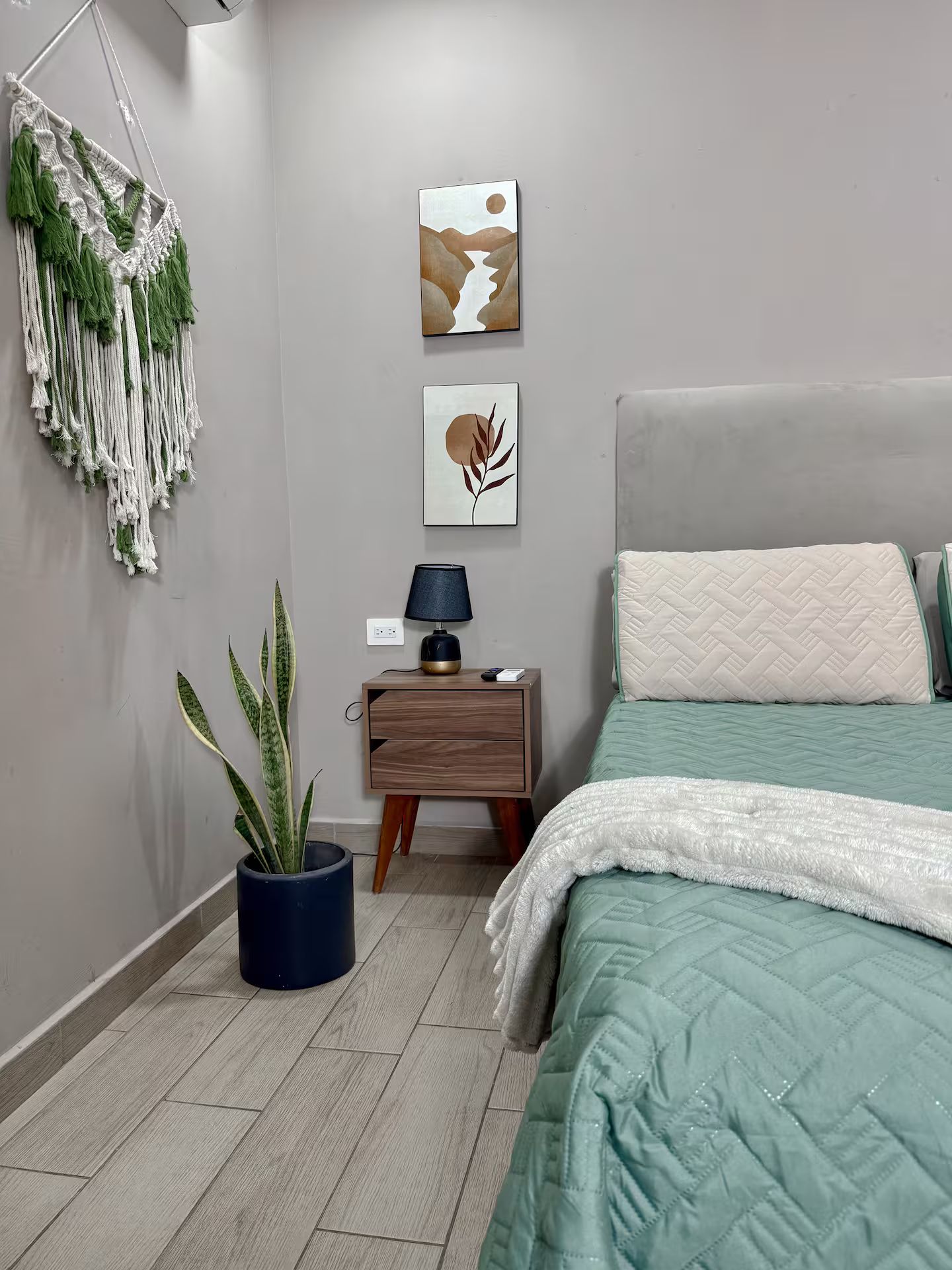 A bedroom with a bed, bedside table, lamp, wall art, macrame wall hanging, and a potted plant.