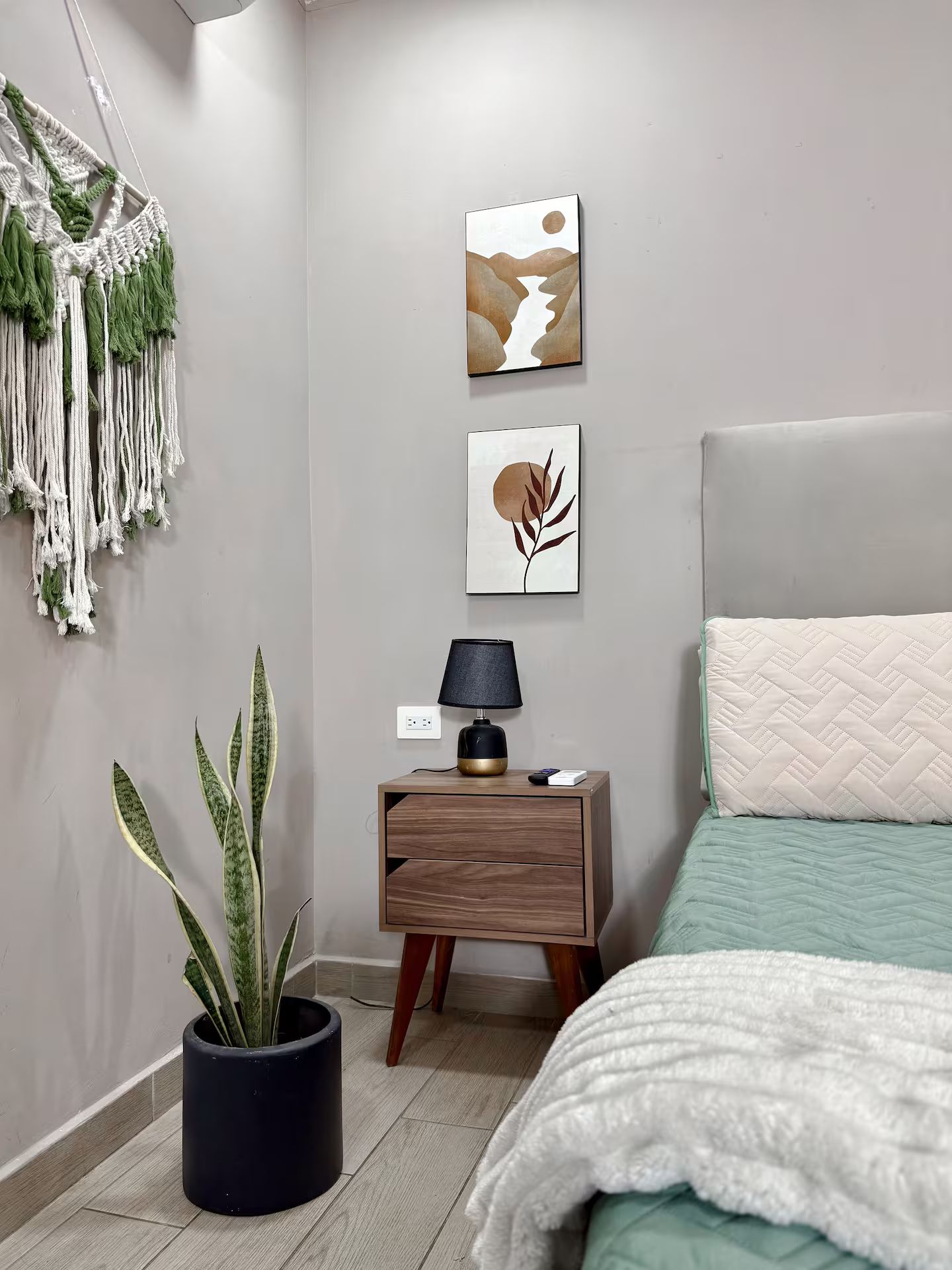 Un dormitorio con cama, mesita de noche, lámpara, decoración en la pared, un colgador de macramé y una planta en maceta.