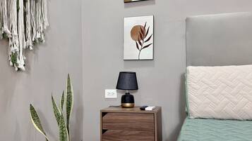 Un dormitorio con cama, mesita de noche, lámpara, decoración en la pared, un colgador de macramé y una planta en maceta.