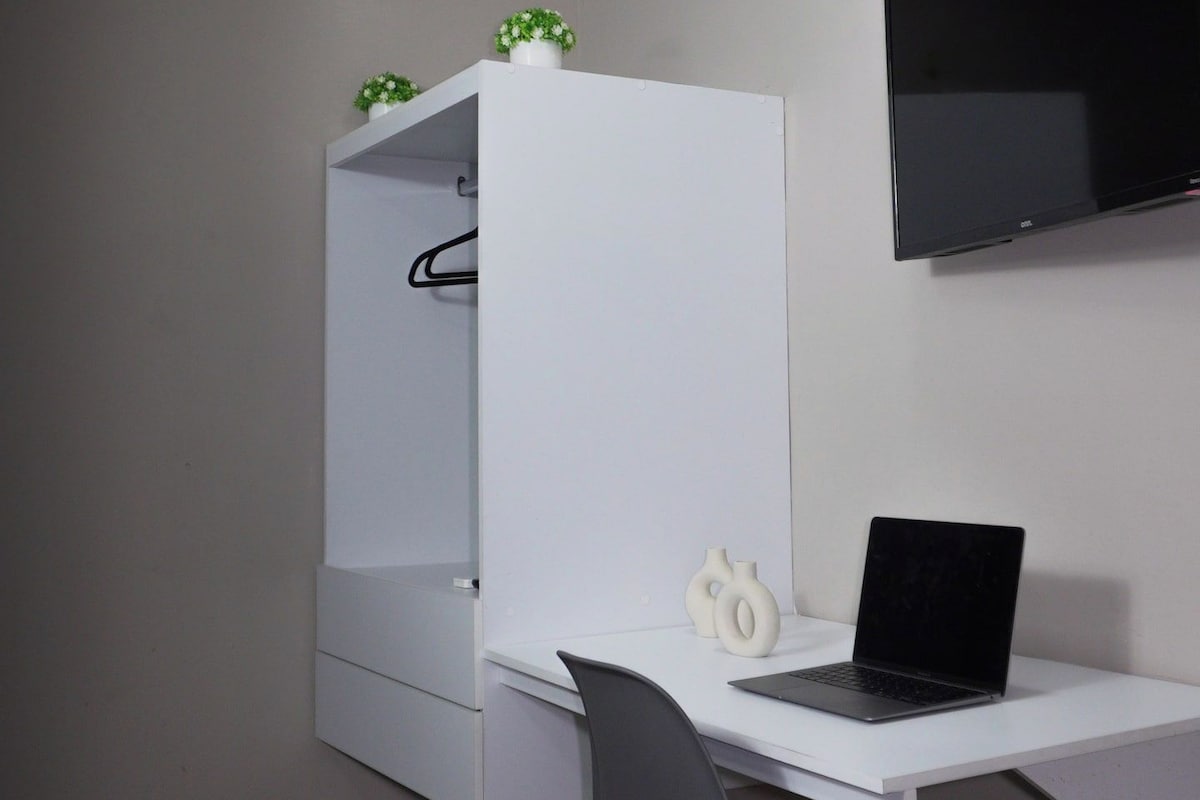 Un dormitorio con cama, un escritorio con computadora portátil, un armario y un televisor montado en la pared.