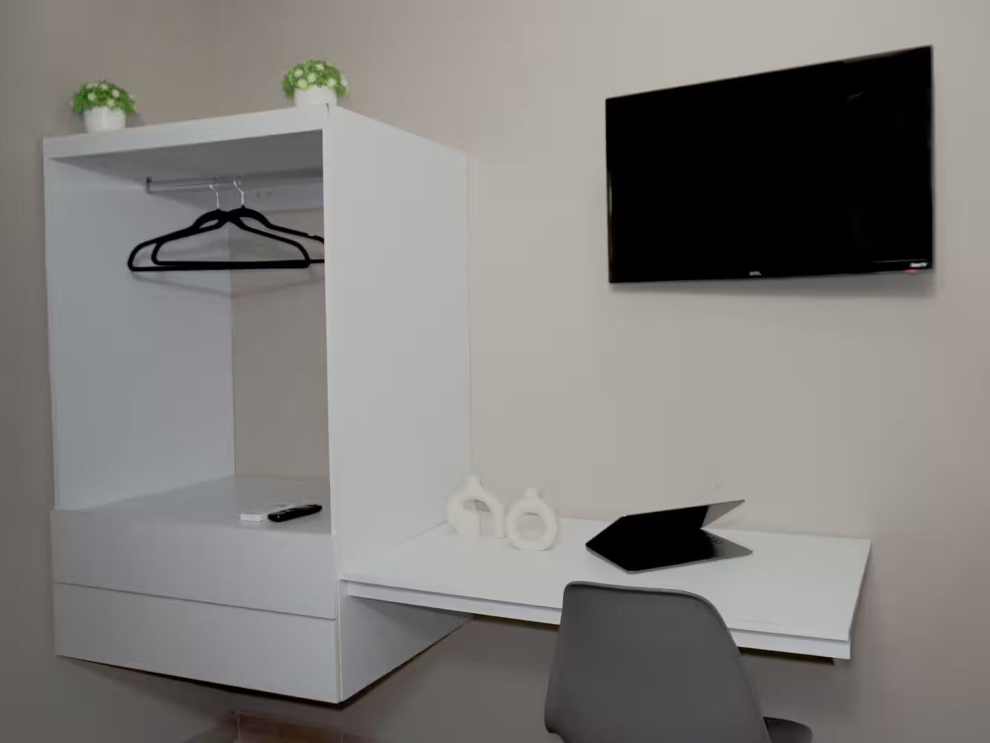 A modern room with a white desk, a black chair, a TV mounted on the wall, and a hanging garment on a rod.