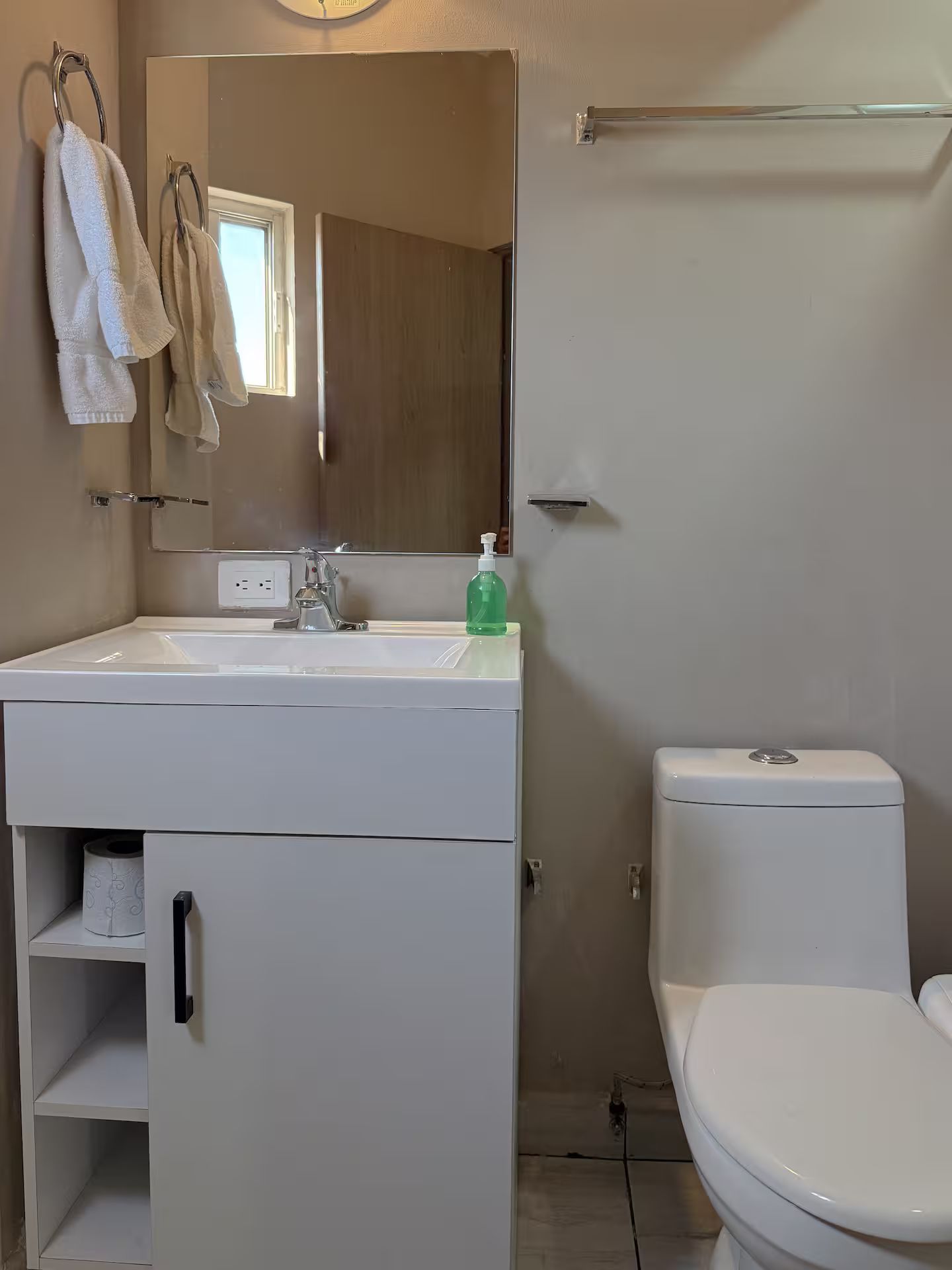 Un baño con lavamanos blanco, espejo y retrete. Hay un perchero con toallas, un dispensador de jabón y un estante con rollo de papel higiénico.