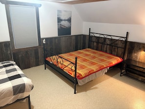 A single bed with a metal frame and a patterned bedspread, a window with blinds, and a wall-mounted picture.
