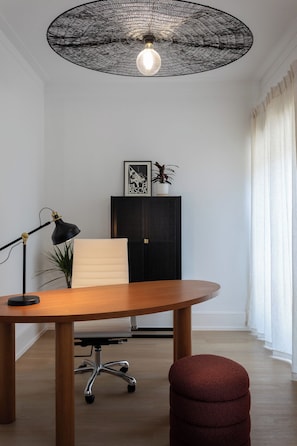 A modern office space with a round wooden table, a black desk lamp, a white office chair, a red ottoman, a black cabinet with a plant, and a unique ceiling light fixture. - Stylish Central Aveiro Stay | 3BR + Office (Aveiro)