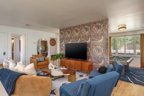 A modern living room with a flat-screen TV, a pool table, and a large window with blinds. - Heatwave HQ-Sport Court, Gym, Pool/SPA, Speakeasy (Scottsdale)