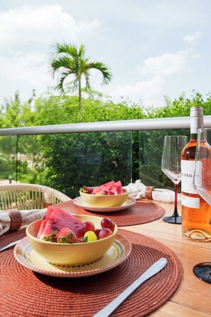 Ein Tisch mit zwei Fruchtschalen, einer Weinflasche und einem Glas auf einem Balkon mit Blick auf Grünflächen.