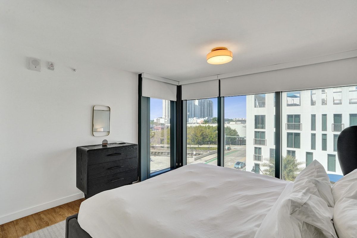 A modern bedroom with a large bed, a nightstand, a mirror, and a view of the city through large windows.