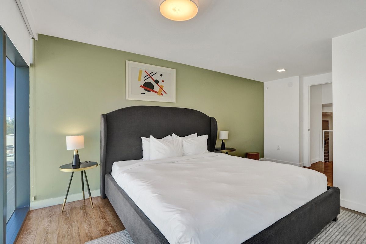 A modern hotel room with a large bed, a side table with a lamp, a framed abstract artwork on the wall, and a view through a glass door.