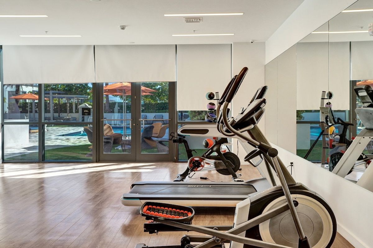 A modern gym with cardio equipment, large windows, and a view of outdoor seating.