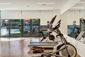Ein modernes Fitnessstudio mit Herz-Kreislauf-Geräten, großen Fenstern und Blick auf eine Außenbestuhlung.