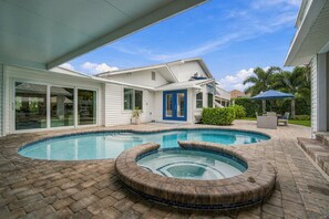 A residential swimming pool with a hot tub, surrounded by a brick patio, and a house with large windows and a blue door. - Coastal Paradise | Waterfront, Heated Pool, Hot Tub & Dock Near Bonita Beach (Bonita Springs)