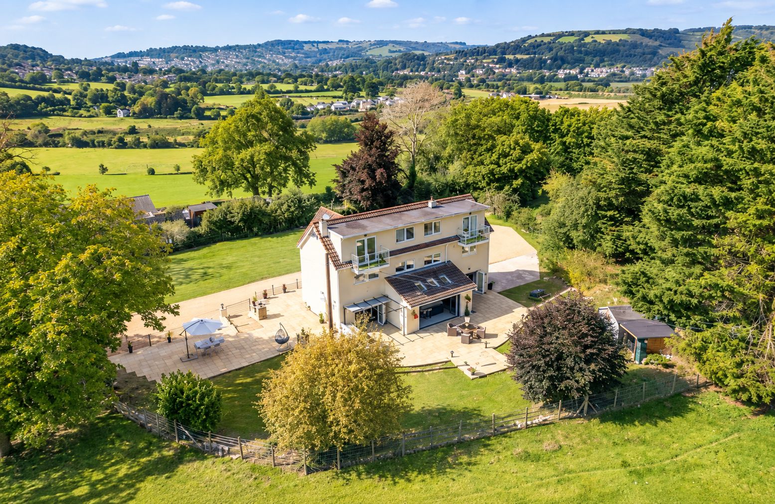 A two-story house with a patio area, surrounded by greenery and a view of a rural landscape.
