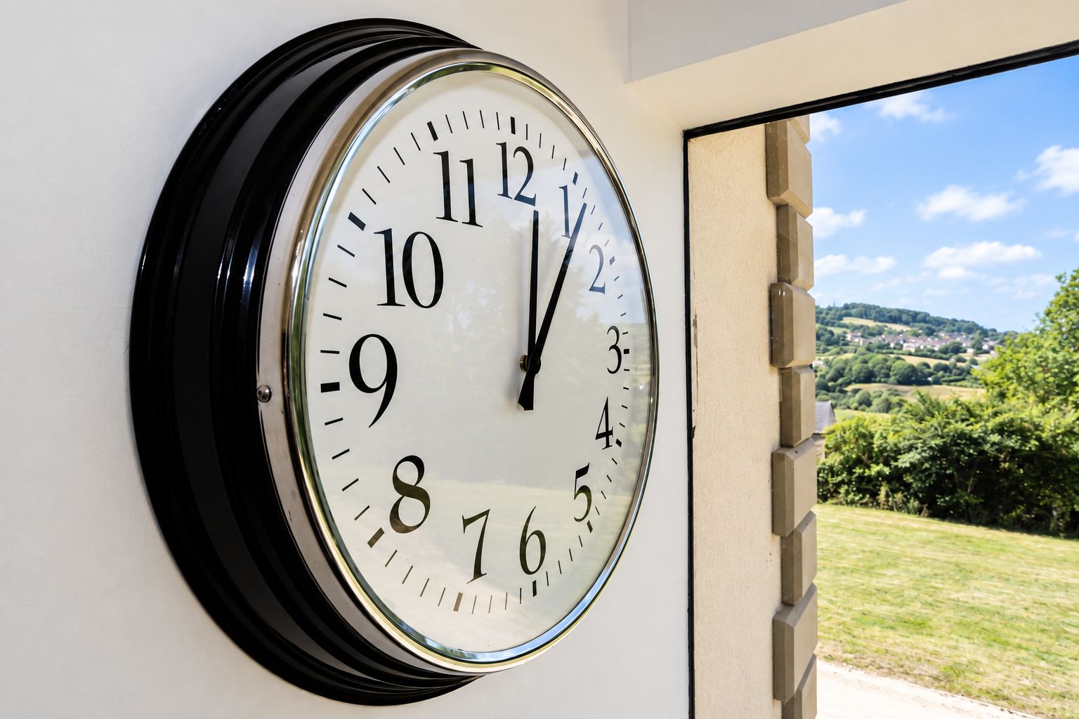 A wall-mounted clock with a clear view of a scenic landscape through an open door.