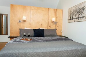 Ein Schlafzimmer mit einem hölzernen Kopfteil, zwei Nachttischlampen, einem Bett mit grauer Tagesdecke und einem Tablett mit zwei Gläsern Rotwein.