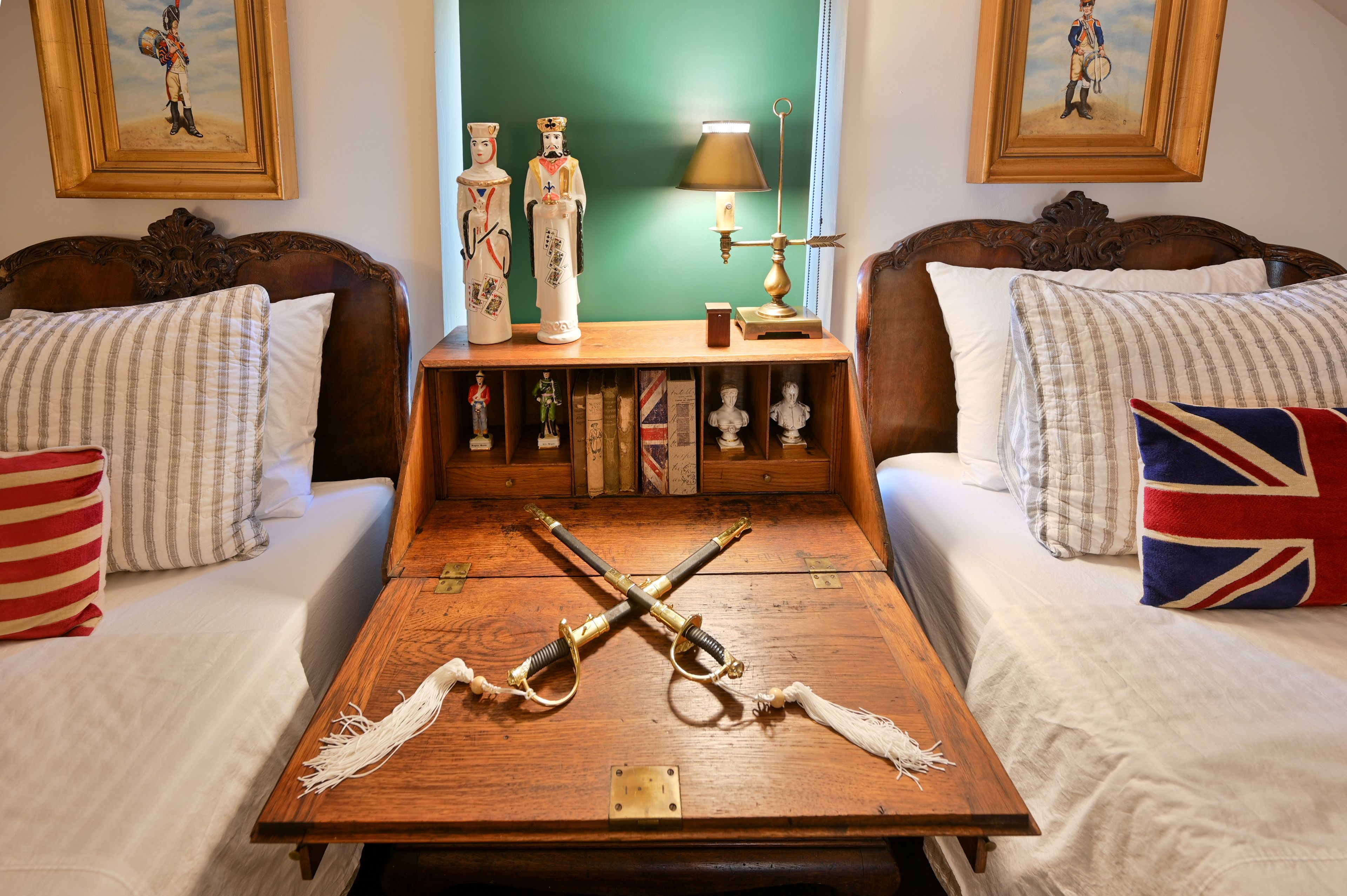 Une chambre avec deux lits, une table en bois sur laquelle sont posés une épée et une lampe, et un mur vert orné de tableaux encadrés.