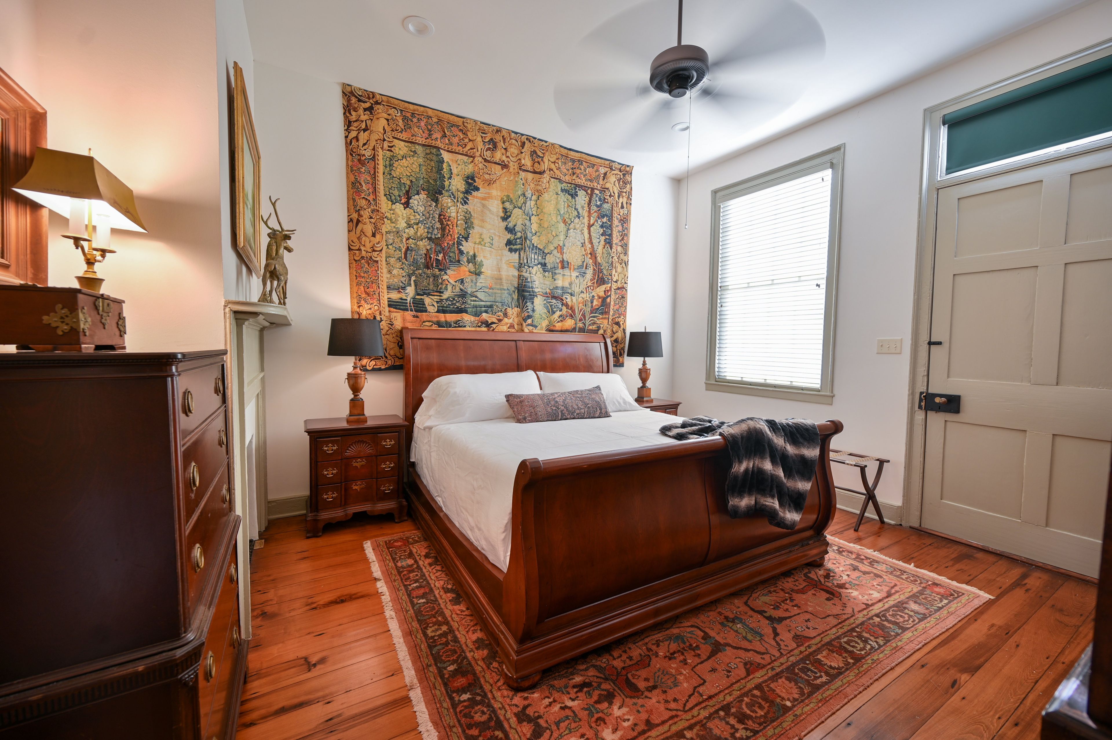 A bedroom with a large bed, a ceiling fan, a tapestry, and a wooden dresser.