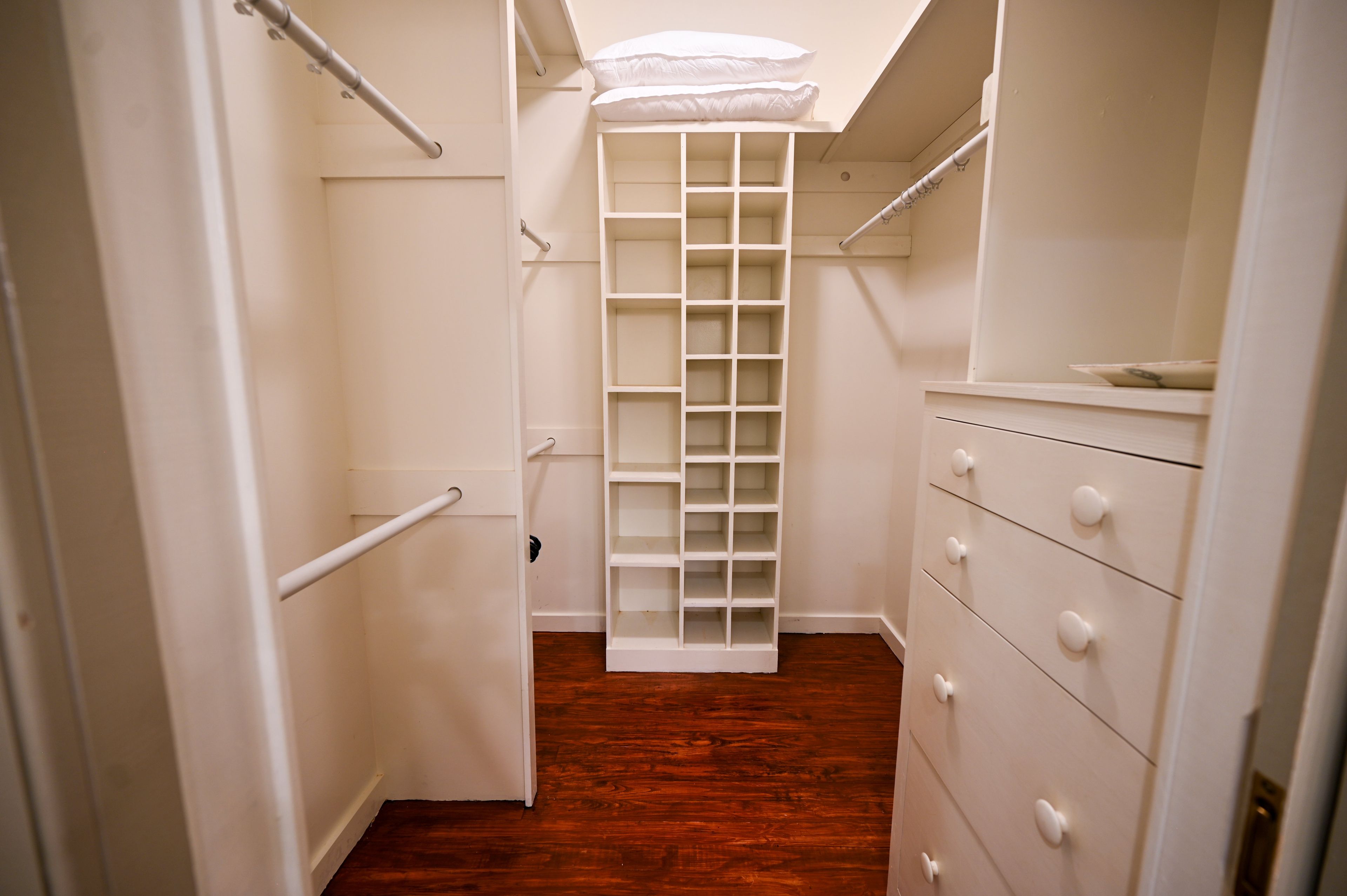 A narrow walk-through closet with wooden flooring, white storage units, and a white ceiling with a skylight.