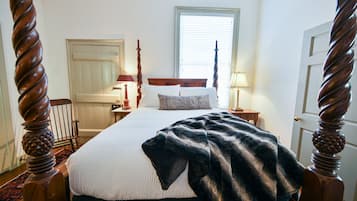 A bedroom with a four-poster bed, a window with blinds, a bedside table with a lamp, and a door.
