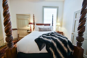 A bedroom with a four-poster bed, a window with blinds, a bedside table with a lamp, and a door.