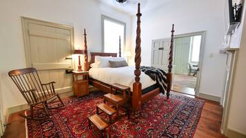 A bedroom with a four-poster bed, a rocking chair, a nightstand, a lamp, and a large patterned rug.