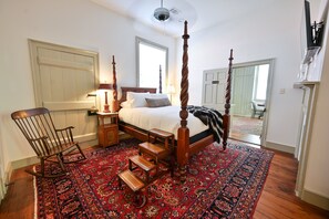 A bedroom with a four-poster bed, a rocking chair, a nightstand, a lamp, and a large patterned rug.