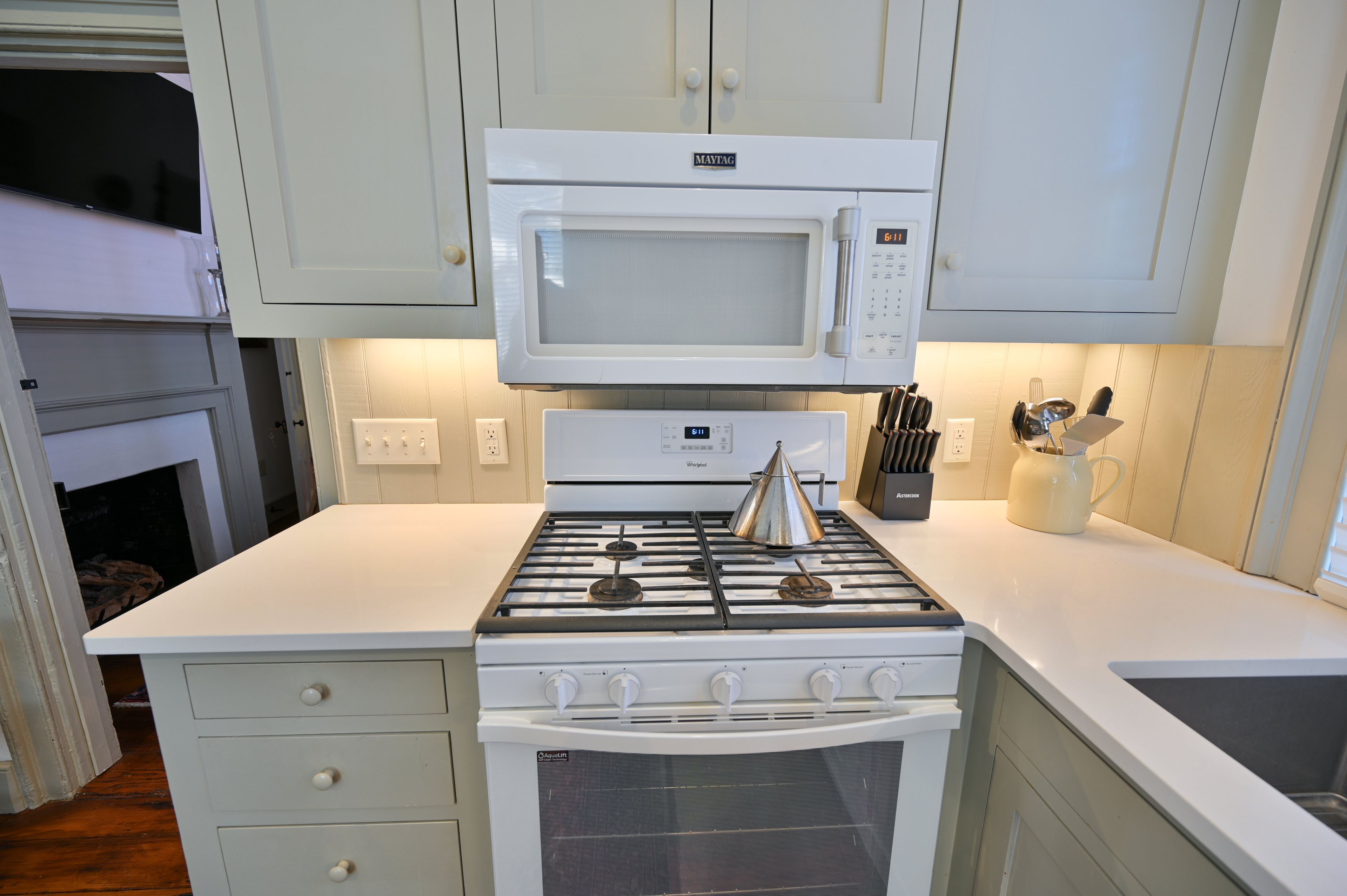 Een kleine keuken met een witte magnetron, fornuis en oven. Boven het fornuis zijn kastjes en er is een klein aanrecht.