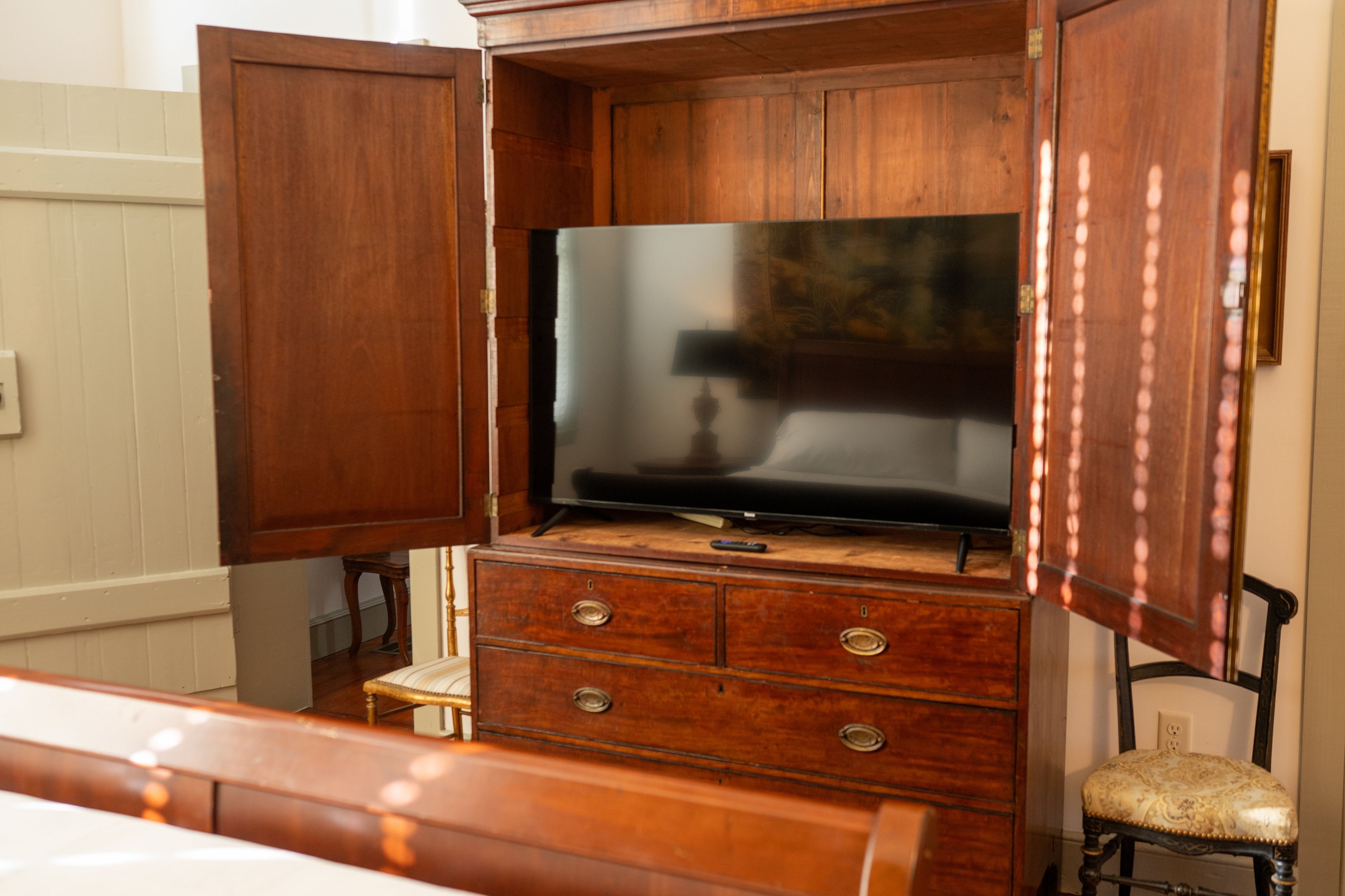 A wooden cabinet with a flat-screen TV on top, a small chair, and a bedside table.