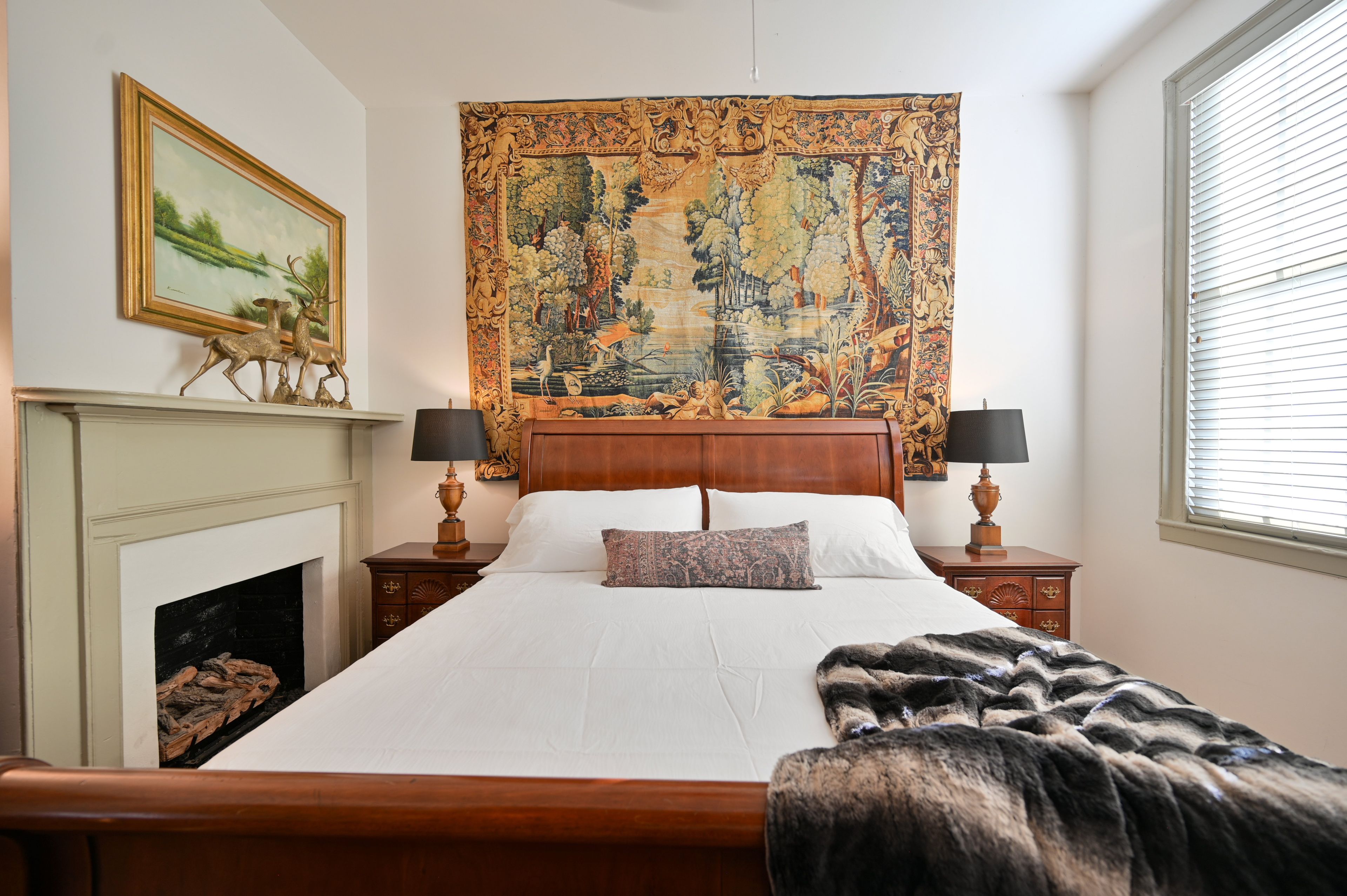 A bedroom with a large bed, a tapestry, a fireplace, and a window with blinds.