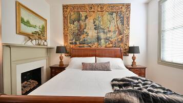 A bedroom with a large bed, a tapestry, a fireplace, and a window with blinds.