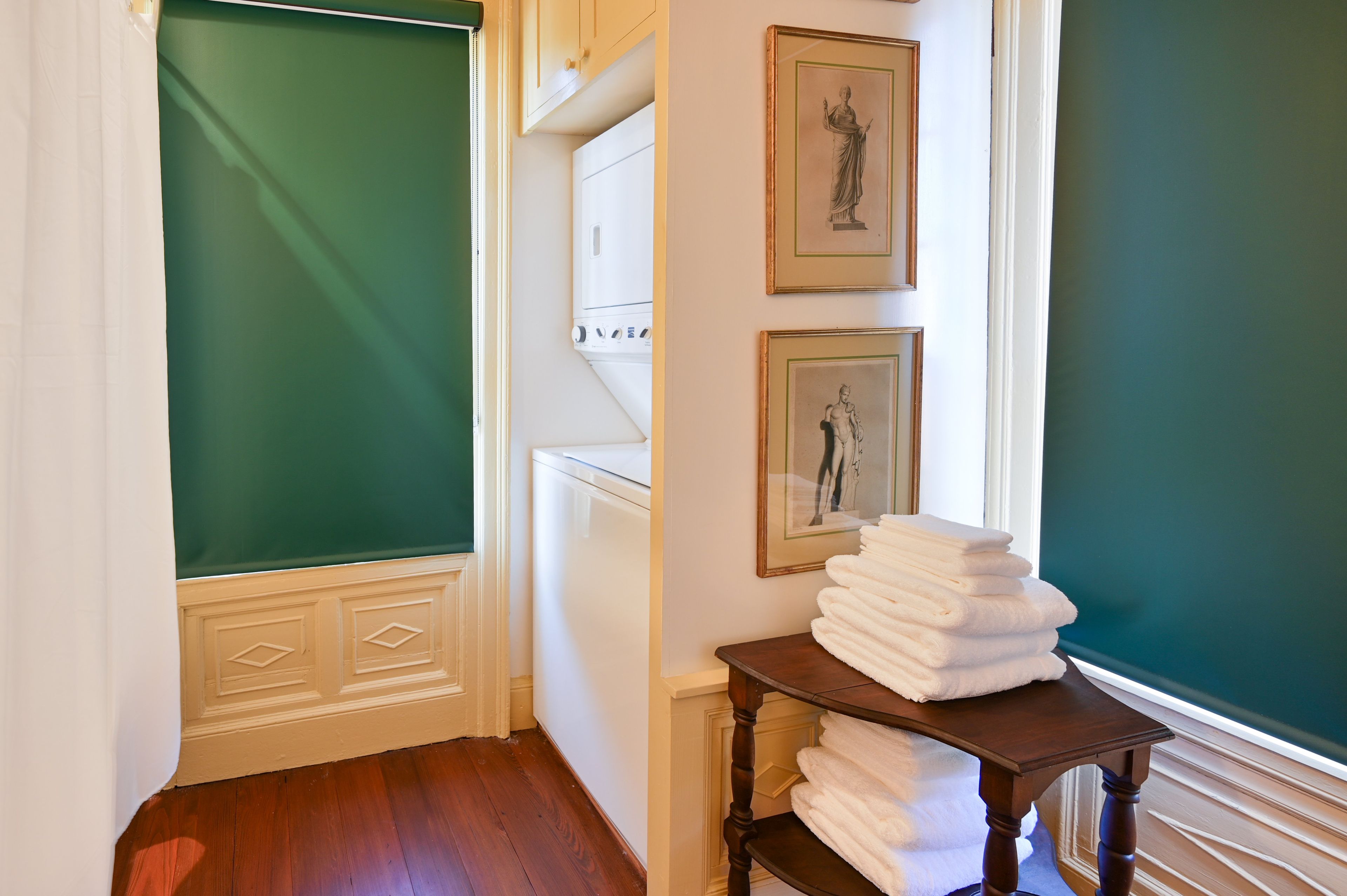 Un pasillo con pared verde, suelo de madera, puerta blanca y una pila de toallas blancas sobre una mesa de madera.