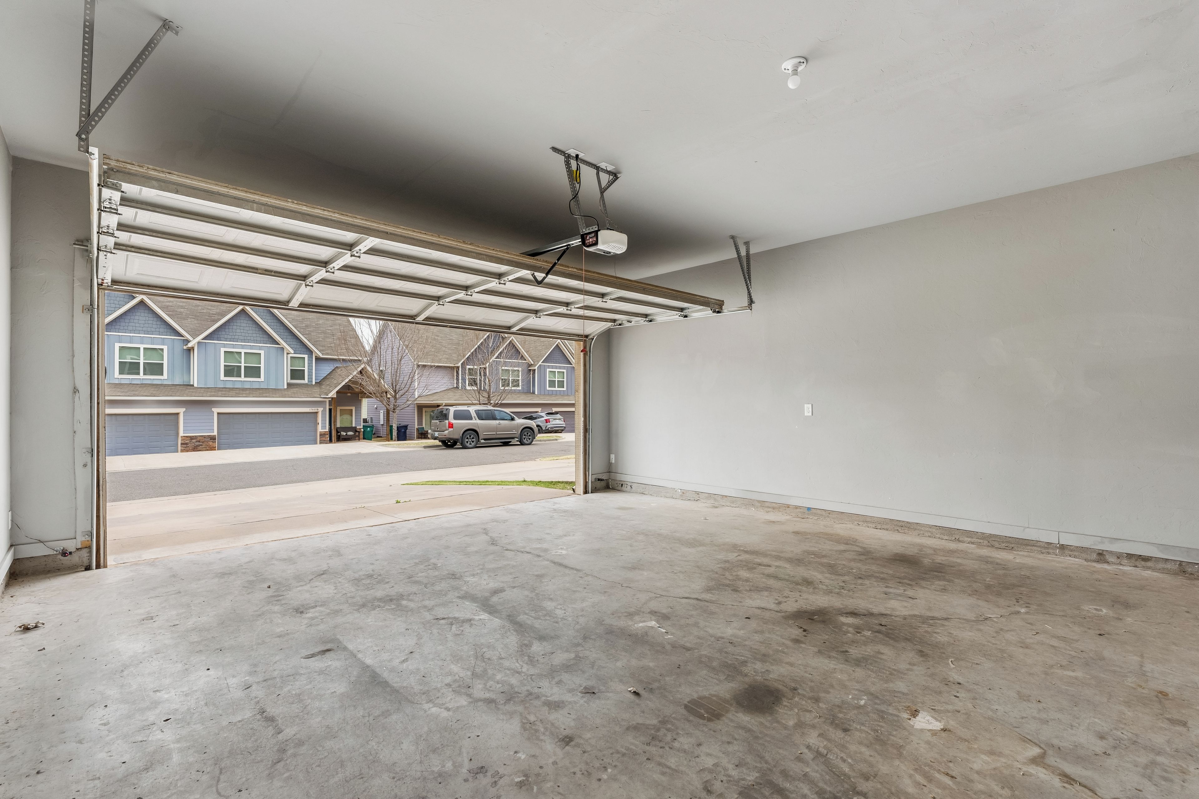 Un garage avec un sol en béton, une porte de garage en métal et une vue sur une rue résidentielle bordée de maisons et de voitures garées.