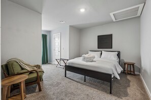 Ein Schlafzimmer mit einem Bett, einem Sessel, einem kleinen Tisch, einer Tür und einem an der Wand hängenden Bild.
