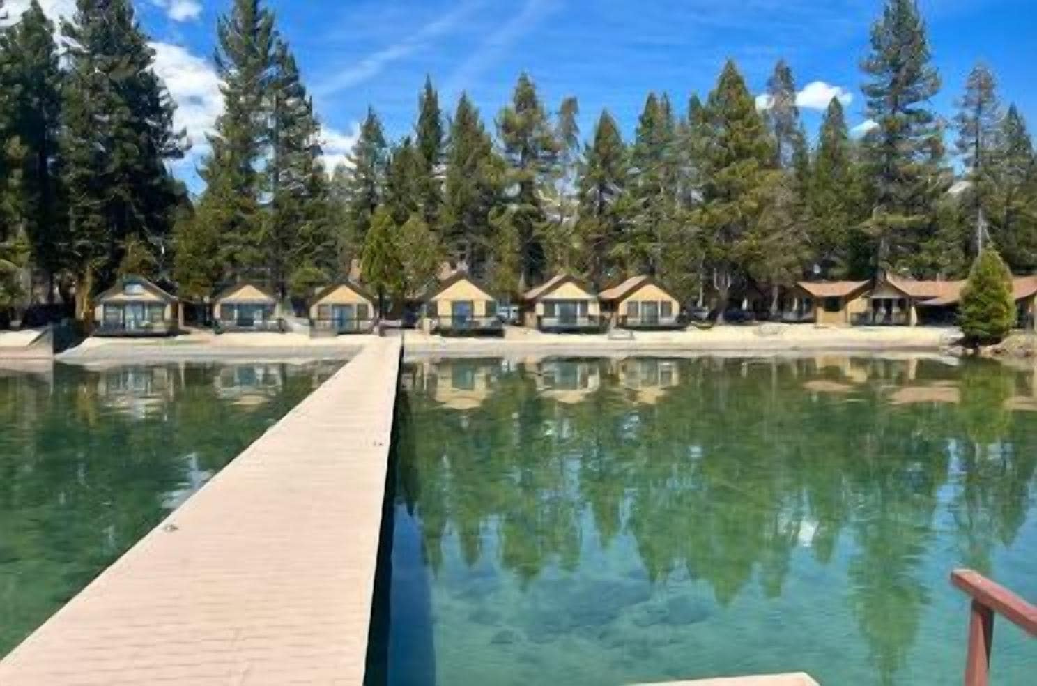A clear swimming pool with a wooden deck, surrounded by forested area and lodge-style buildings.
