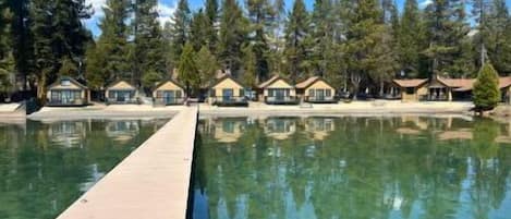 A clear swimming pool with a wooden deck, surrounded by forested area and lodge-style buildings.