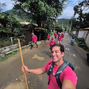 A group of people on a dirt road with a bamboo pole, some carrying backpacks, and a small structure in the background.