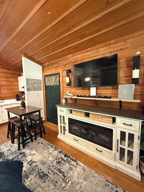 A cozy cabin interior with a wooden ceiling, a flat-screen TV mounted on the wall, a dining table with chairs, and a fireplace. - Captain's Cabin at Elk Meadows Cabins (Forks)