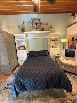 A bedroom with a large bed, built-in shelving, a chair, and a wall-mounted decorative piece. - Captain's Cabin at Elk Meadows Cabins (Forks)
