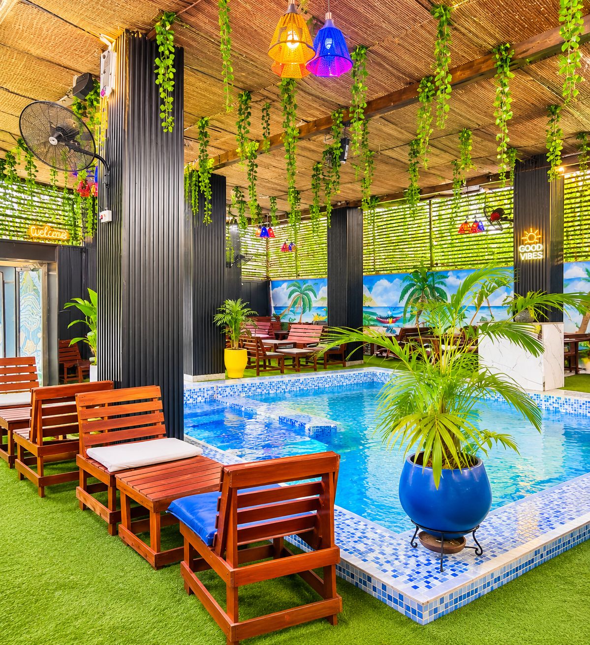 An indoor pool area with wooden chairs, a blue mosaic pool, hanging plants, and a tropical mural.